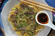 A plate of Crispy fried noodles with pork and green vegetables serve with Mei's Homemade Chiu Chow Chilli Oil. A ceramic spoon filled with chilli oil and chilli crisps, a pair of chopsticks.